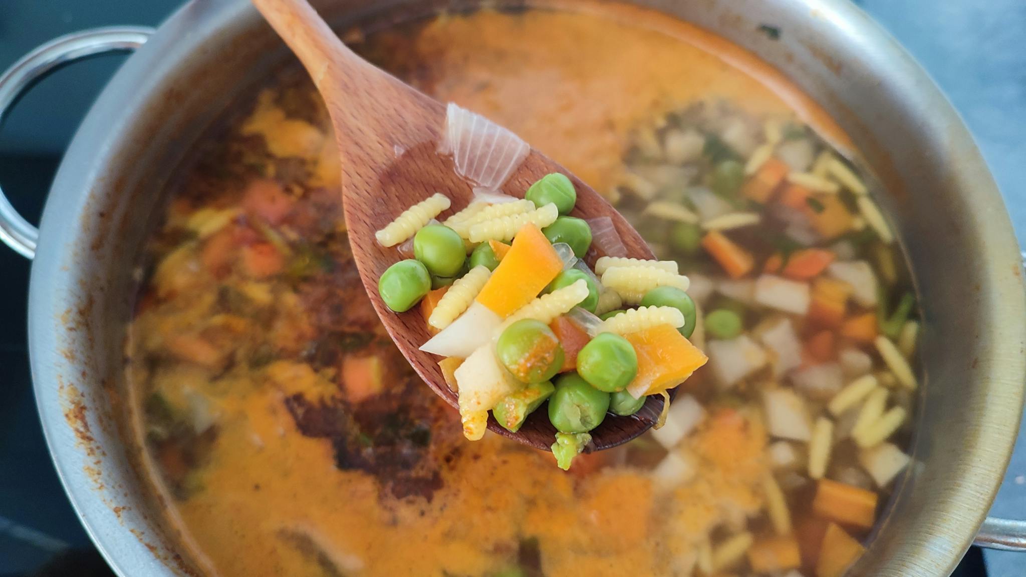 Close-up of a wooden spoon holding fresh vegetables over simmering soup.